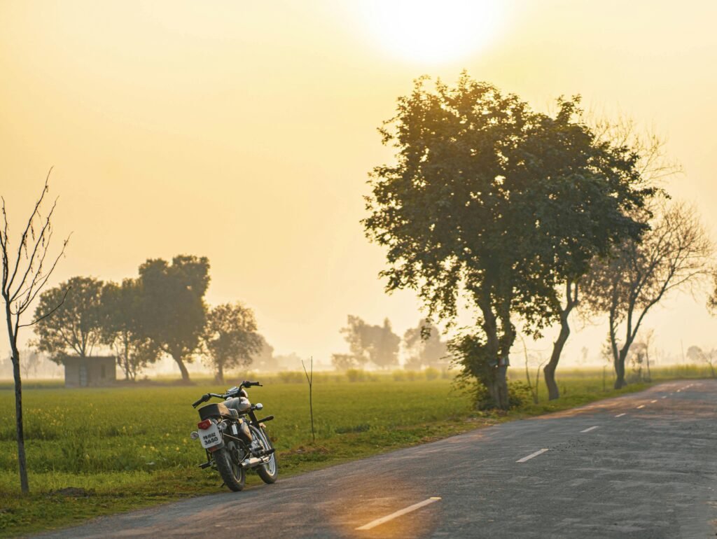 Serene image of a motorcycle parked on a rural road at sunrise in India with trees and fields in the background.